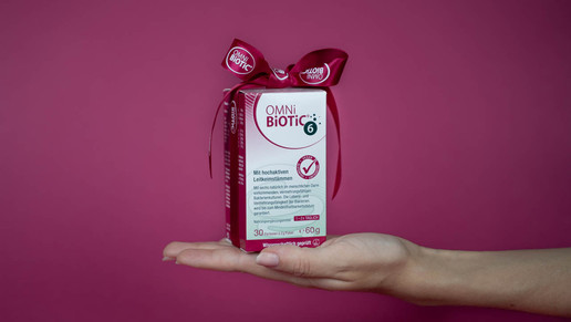 Hand hält eine Packung OMNi-BiOTiC 6 mit Geschenk-Schleife vor pinkem Hintergrund – Nahrungsergänzung mit hochaktiven Leitkeimstämmen für die Darmgesundheit.