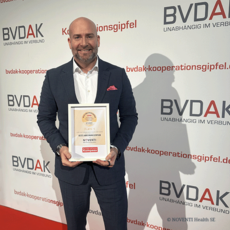 Mark Böhm, Vorstandsvorsitzender NOVENTI Health SE, vor einer Fotowand mit dem Schriftzug 'BVDAK Kooperationsgipfel'. Er hält eine gerahmte Urkunde mit dem NOVENTI Logo sowie dem Titel 'Deutschlands bestes Abrechnungszentrum 2026' in beiden Händen.