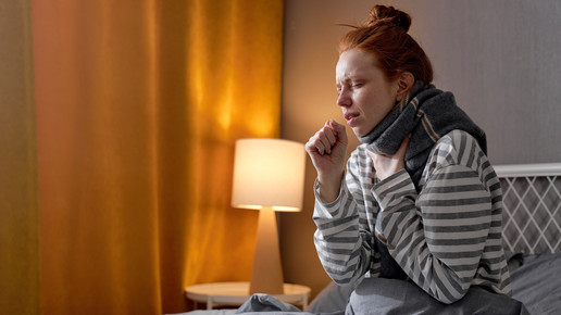 Foto: Eine Person sitzt zugedeckt mit Schal im Bett und hustet