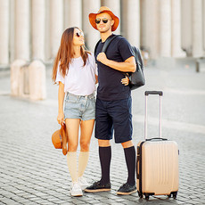 Die Abbildung zeigt ein junges Pärchen. Sie ist angezogen mit einer kurzen Jeans und einen weißen T-Shirt, hat einen Sommerhut in der Hand. Er ist ganz in dunkelblau angezogen, T-Shirt, kurze Hose. Sie tragen beide Reise Kniestrümpfe, vor ihnen steht ein Koffertrolley.