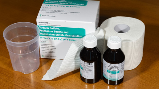 Auf einem Tisch stehen ein Plastikmessbecher, eine Packung mit Abführmitteln und eine Rolle Toilettenpapier.