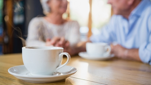 Foto: Zwei ältere Personen im Hintergrund, im Vordergrund fokussiert eine Tasse Tee/Kaffee