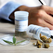 Foto: Zwei Abgabegefäße mit Medizinalcannabis auf einem Tisch, eine schreibende Person im Kittel im Hintergrund.