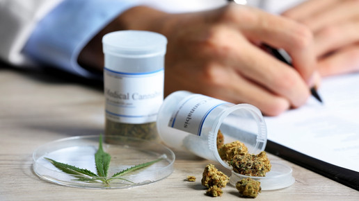 Foto: Zwei Abgabegefäße mit Medizinalcannabis auf einem Tisch, eine schreibende Person im Kittel im Hintergrund.