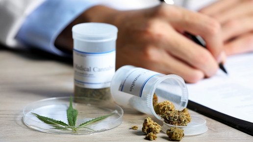 Foto: Zwei Abgabegefäße mit Medizinalcannabis auf einem Tisch, eine schreibende Person im Kittel im Hintergrund.