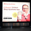 Das Foto zeigt ein Werbeplakat des Versenders ShopApotheke. TV-Moderator Günther Jauch hält ein Handy hoch, auf dem die ShopApotheke-App geöffnet ist. Er hält eine Gesundheitskarte von hinten an das Gerät. Auf dem Plakat steht: „E-Rezept? Einfach ShopApotheke-App. Jetzt herunterladen und Wege sparen.“