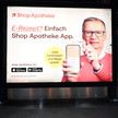 Das Foto zeigt ein Werbeplakat des Versenders ShopApotheke. TV-Moderator Günther Jauch hält ein Handy hoch, auf dem die ShopApotheke-App geöffnet ist. Er hält eine Gesundheitskarte von hinten an das Gerät. Auf dem Plakat steht: „E-Rezept? Einfach ShopApotheke-App. Jetzt herunterladen und Wege sparen.“