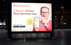Das Foto zeigt ein Werbeplakat des Versenders ShopApotheke. TV-Moderator Günther Jauch hält ein Handy hoch, auf dem die ShopApotheke-App geöffnet ist. Er hält eine Gesundheitskarte von hinten an das Gerät. Auf dem Plakat steht: „E-Rezept? Einfach ShopApotheke-App. Jetzt herunterladen und Wege sparen.“