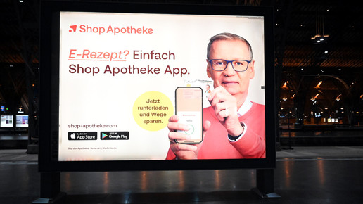 Das Foto zeigt ein Werbeplakat des Versenders ShopApotheke. TV-Moderator Günther Jauch hält ein Handy hoch, auf dem die ShopApotheke-App geöffnet ist. Er hält eine Gesundheitskarte von hinten an das Gerät. Auf dem Plakat steht: „E-Rezept? Einfach ShopApotheke-App. Jetzt herunterladen und Wege sparen.“