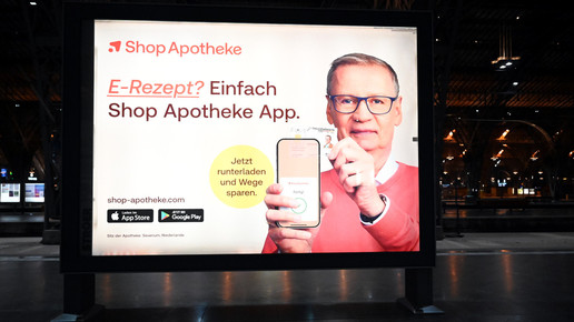 Das Foto zeigt ein Werbeplakat des Versenders ShopApotheke. TV-Moderator Günther Jauch hält ein Handy hoch, auf dem die ShopApotheke-App geöffnet ist. Er hält eine Gesundheitskarte von hinten an das Gerät. Auf dem Plakat steht: „E-Rezept? Einfach ShopApotheke-App. Jetzt herunterladen und Wege sparen.“