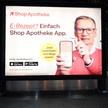 Das Foto zeigt ein Werbeplakat des Versenders ShopApotheke. TV-Moderator Günther Jauch hält ein Handy hoch, auf dem die ShopApotheke-App geöffnet ist. Er hält eine Gesundheitskarte von hinten an das Gerät. Auf dem Plakat steht: „E-Rezept? Einfach ShopApotheke-App. Jetzt herunterladen und Wege sparen.“