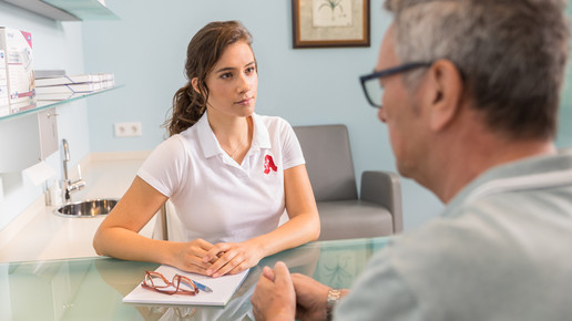 Eine Apothekenangestellte im weißen Shirt mit rotem Apotheken-A sitzt gegenüber von einem Kunden am Tisch. Mehr dazu im Artikel.