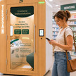 Frau am Handy steht vor der Doctorbox-Kabine in einer Apotheke