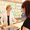 Foto: Eine Person im weißen Kittel in der Apotheke bearbeitet ein Papierrezept vor einer Kundin.