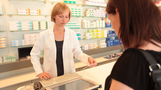 Foto: Eine Person im weißen Kittel in der Apotheke bearbeitet ein Papierrezept vor einer Kundin.