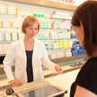 Foto: Eine Person im weißen Kittel in der Apotheke bearbeitet ein Papierrezept vor einer Kundin.