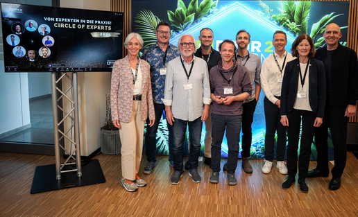 „Gruppenfoto von neun Personen auf einer Konferenz, stehend vor einer beleuchteten Wand; links im Bild steht ein Bildschirm mit der Aufschrift ‚Circle of Experts‘.“