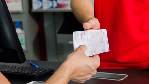 Foto: Eine Person reicht einer anderen Person ein Muster-16-Rezept in einer Apotheke