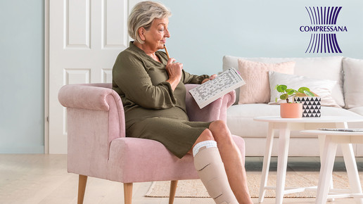 Abgebildet ist eine Dame auf einem Sessel sitzend in einem Wohnzimmer.Sie hat an ihrem rechten Unterschenkel eine SoftCompress Bandagehilfe angelegt und trägt darüber ein medizinisch adaptives Kompressionssystem in der Farbe Beige.