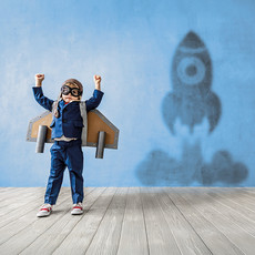 Ein Junge trägt eine Fliegerbrille und Flügel aus Pappe auf dem Rücken. Sein Schatten ist eine startende Rakete.