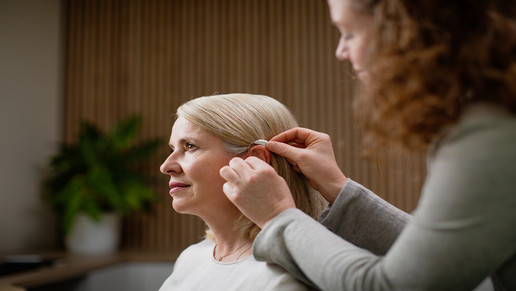 „EINE FACHKRAFT PASST EINER SITZENDEN FRAU EIN HÖRGERÄT HINTER DEM OHR AN.“