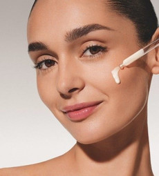 Nahaufnahme einer lächelnden Frau mit zurückgebundenem Haar. Eine Glaspipette trägt einen Tropfen einer weißen, leichten Serum-Textur direkt auf ihre linke Wange auf. Der Hintergrund ist hell und neutral gehalten.