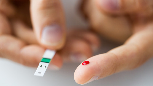 Zwei Hände. Eine hält einen Teststreifen. An der anderen ragt ein Tropfen Blut an der Fingerbeere heraus. Es wird Blutzucker gemessen.