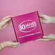 Zwei Hände halten eine pinke Geschenkbox mit der Aufschrift ‚30 Jahre OMNi-BiOTiC – Bei uns ist Forschung zu Hause‘ vor pinkem Hintergrund.