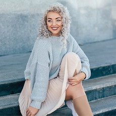 Eine Frau mit grau gelockten langen Haaren, bekleidet mit einem beige farbenen Plissee Rock und einen hellblauen Rippenpullover sitzt auf einer Treppe und lächelt. Sie trägt Kompressionskniestrümpfe in beiger Farbe.