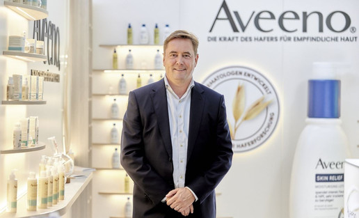 Mann in dunklem Anzug steht vor einem Aveeno® Messestand mit Hautpflegeprodukten und großem Logo 'Aveeno® Die Kraft des Hafers für empfindliche Haut'
