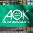 Ein Schild der AOK Krankenkasse hängt an einem Zaun.