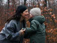 Mutter und Kind im herbstlichen Wald. Sie halten sich an den Händen und stehen sich gegenüber. Beide tragen warme Kleidung.
