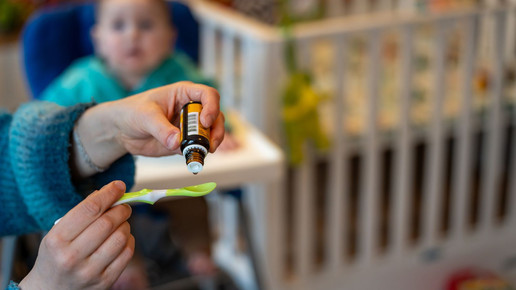 Symbolbild: Eine Person tropft ein flüssiges Vitamin-D-Präparat auf einen Löffel, im Hintergrund sitzt ein Baby.