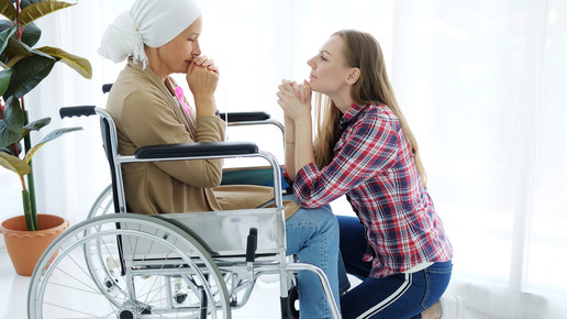 Kranke Frau sitzt im Rollstuhl, vor ihr kniet eine Angehörige.