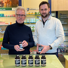 Zwei Männer in einer Apotheke zeigen Gelenktabletten für Hunde; fünf schwarze Dosen stehen auf dem Tresen, Regale mit Produkten im Hintergrund.