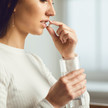 Foto: Eine Frau im Profil hält eine Tablette vor ihre Lippen und hält ein Glas Wasser in ihrer Hand.