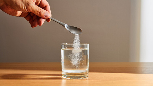 Eine Hand lässt über einen Löffel weißes Pulver in ein Wasserglas rieseln