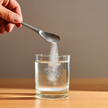 Eine Hand lässt über einen Löffel weißes Pulver in ein Wasserglas rieseln