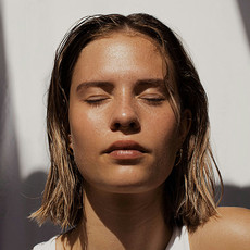 Eine junge Frau mit nassen, kinnlangen Haaren steht mit geschlossenen Augen in der Sonne. Sie trägt ein weißes Oberteil, und ihr Gesicht wird von hellem Sonnenlicht beleuchtet. Der Hintergrund ist unscharf, mit hellen und dunklen Stoffbahnen.