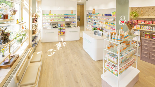Ein heller, modern gestalteter Verkaufsraum einer Apotheke mit hellem Laminatboden in Holzoptik. Links befinden sich große Fensterfronten mit Pflanzen und einer hölzernen Sitzbank für Kunden. Im Hintergrund stehen zwei weiße Verkaufstresen mit geriffelter Front. Die Wände sind mit hellen Holzregalen bestückt, in denen Medikamente und Pflegeprodukte ordentlich sortiert sind. Ein dezentes Logo – eine stilisierte weiße Figur mit einem roten Herz – ist an einer grünen Wandfläche rechts zu sehen. Die Atmosphäre ist lichtdurchflutet und freundlich.