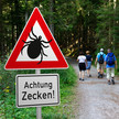 Symbolbild: Ein Warnschild mit der Aufschrift „Achtung Zecken!“ steht gut sichtbar am Rande eines Waldwegs; im Hintergrund ist eine Familie beim Spaziergang zu sehen. 