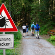 Faktencheck FSME: Deutschland als Endemiegebiet Symbolbild: Ein Warnschild mit der Aufschrift „Achtung Zecken!“ steht gut sichtbar am Rande eines Waldwegs; im Hintergrund ist eine Familie beim Spaziergang zu sehen.