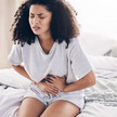 Symbolbild: Frau sitzt auf einem Bett und hält sich mit schmerzverzerrtem Gesicht den Unterleib