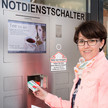 Eine Dame steht vor der DACOS Abholstation mit einem offenen Fach und Medikamenten in der Hand.