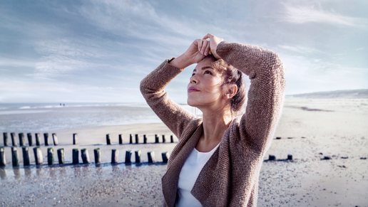 Eine Frau mit hochgesteckten Haaren steht an einem weiten Sandstrand. Sie trägt eine beige Strickjacke über einem weißen Oberteil und hält die Hände über dem Kopf. Der Himmel ist leicht bewölkt, das Licht wirkt kühl und klar, im Hintergrund das Meer.