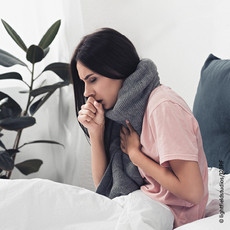 Junge Frau mit dunklen Haaren, die ein T-Shirt trägt und einen dicken Schal um den Hals, sitzt im Bett und hustet.