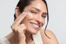 Nahaufnahme einer Frau mit dunklen, zurückgebundenen Haaren, die mit zwei Fingern Creme unter ihr rechtes Auge aufträgt. Sie lächelt freundlich, trägt ein ärmelloses, helles Oberteil, und der Hintergrund ist neutral hell.