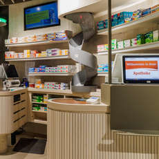 Moderner Verkaufsraum einer Apotheke mit Arzneimittelregalen, Preisauszeichnungen und digitalen Bildschirmen.