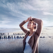 Eine Frau mit hochgesteckten Haaren steht an einem weiten Sandstrand. Sie trägt eine beige Strickjacke über einem weißen Oberteil und hält die Hände über dem Kopf. Der Himmel ist leicht bewölkt, das Licht wirkt kühl und klar, im Hintergrund das Meer.