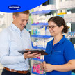 Ein Mann und eine Frau in Arbeitskleidung stehen in einer Apotheke oder einem Sanitätshaus und schauen gemeinsam auf ein Tablet. Beide lächeln und wirken im Gespräch vertieft.
